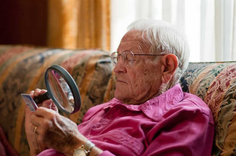 Elderly old man using magnifying glass to read fine print on object with strong light over shoulder while wearing glasses. Photo: Getty