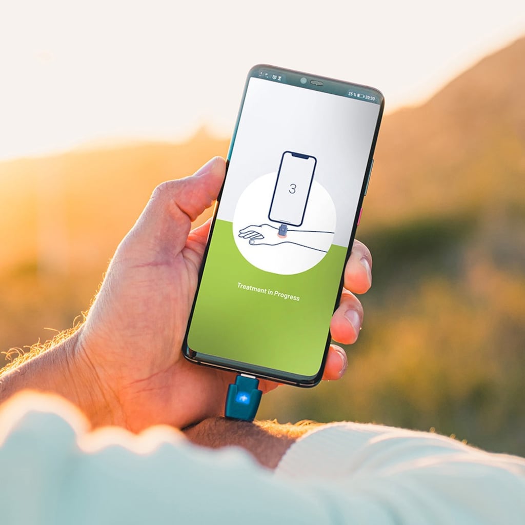 a smartphone with a small blue dongle plugged into the bottom, and a screen displaying a treatment in progress