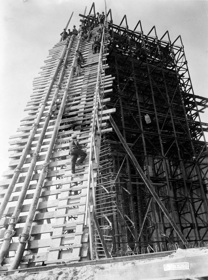 A prisoner climbs scaffolding. The three pipes to his left pump cement to the waiting prisoners above