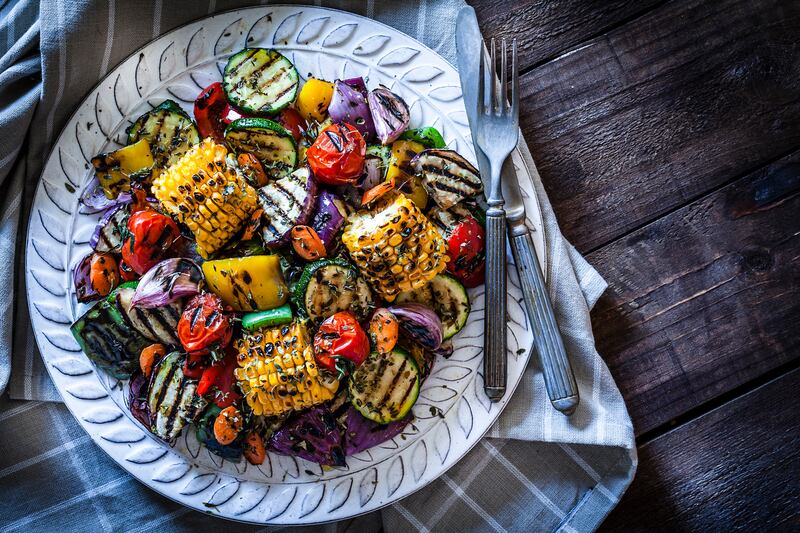 Close-up view of a grilled vegetables plate shot from above on rustic wooden kitchen table. The plate is on a gray textile napkin at the left of an horizontal frame and a fork and knife are at the right of the plate. DSRL studio photo taken with Canon EOS 5D Mk II and Canon EF 100mm f/2.8L Macro IS USM