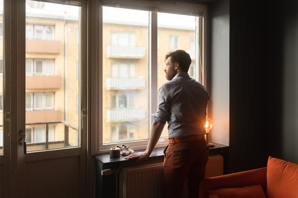 A man at home alone in his apartment. He is standing by the window, looking out on the city.