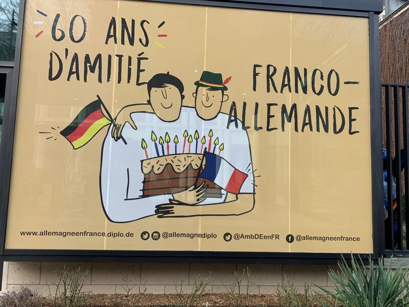 The mural outside the German embassy in Paris shows a Frenchman in a beret embracing a German wearing a Tyrolean hat. The two hold their national flags and a birthday cake with candles. It reads: “60 Years of Franco-German Friendship.”