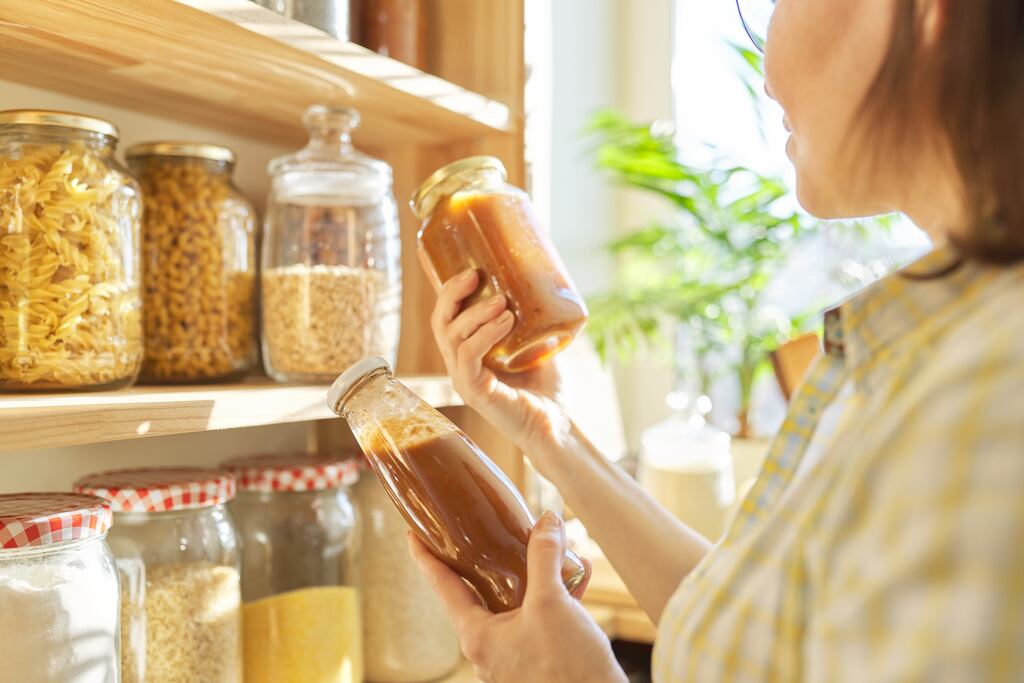 Woman holding bottle with ketchup, picking food from storage cabinet in kitchen, storage with wooden shelves