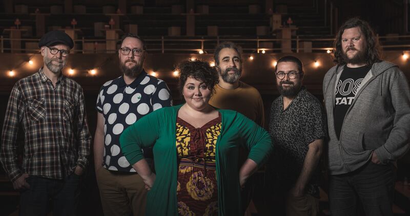 Singer, Ríoghnach Connolly stands wearing a green cardigan and brown and yellow dress, in front of her 5 Honeyfeet band mates