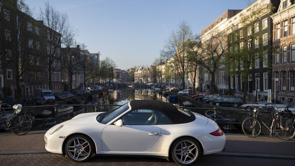 A white Porsche AG automobile sits parked on a bridge over a canal in Amsterdam, Netherlands.