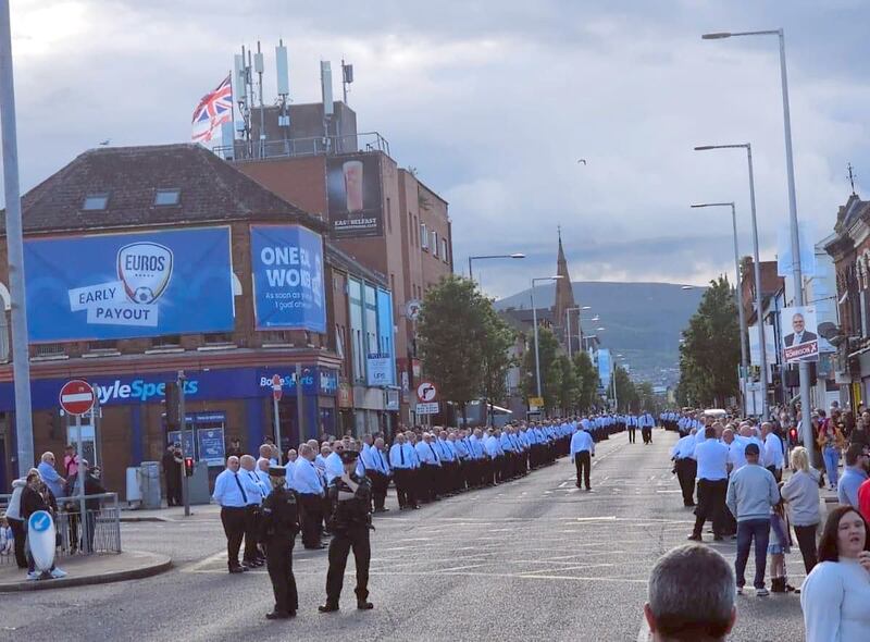 Men in white shirts and black ties line the route of a memorial parade on the Newtownards Road in June 2024 