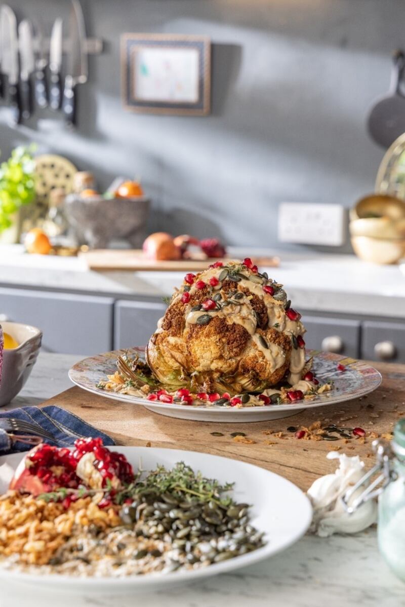 Roasted whole cauliflower with plate of crispy onion pieces and toasted pumpkin, linseed and sesame seeds studded with pomegranate seeds