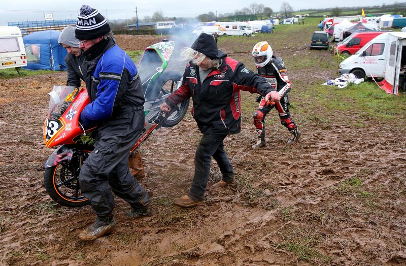 Sam Dunlop trudges through the muddy paddock at the 2016 Mid Antrim 150 as his father, Jim, helps carry his son’s race bike to the start line.  Photograph: Stephen Davison
