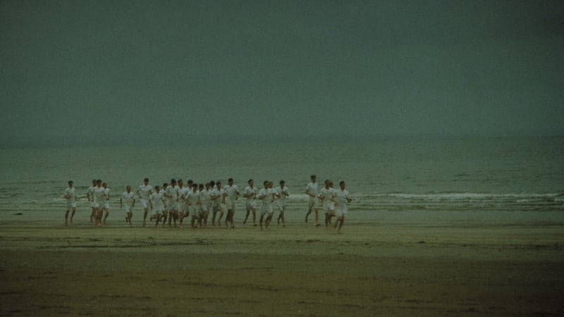 A slow-motion shot depicting 1924 Olympians running barefoot along a wide, open beach, sand splashing from the shallow shore on to their all-white uniforms