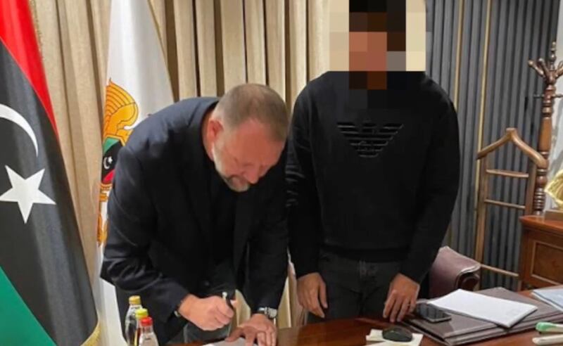 Danny Cluskey signing papers at a desk in an office with the flag of Khalifa Haftar’s Libyan faction behind him