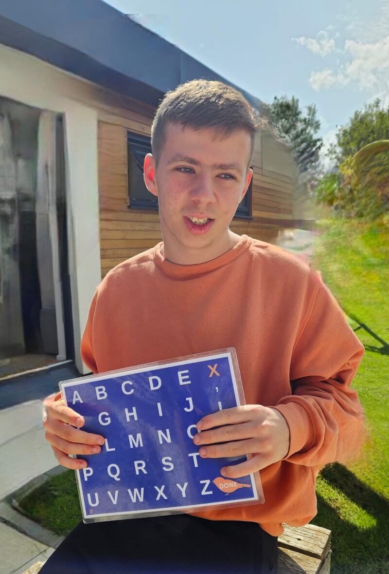 Caoimh holds up a spell-to-communicate (S2C) letterboard. Regular practice in controlling his arm movements so that he can point accurately at the letterboard, as well as the assistance of a trained S2C communication and regulation partner, are what enable Caoimh to discuss his medical symptoms with his doctors
